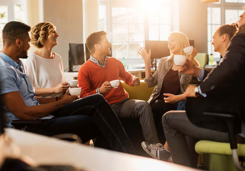 Drei Frauen un ddrei Männer sitzen im Büro in einem Stuhkreisen, heben ihre Kaffeetasen und haben Spaß.