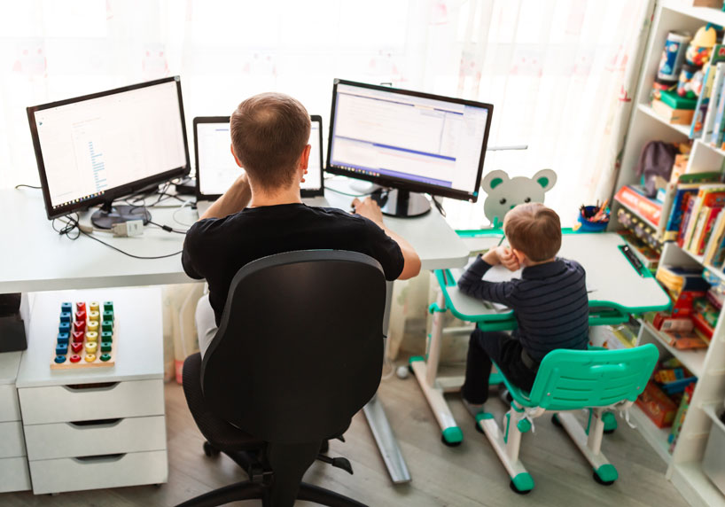 Mann arbeitet im Homeoffice und neben ihm sitzt sein Sohn am Maltisch.